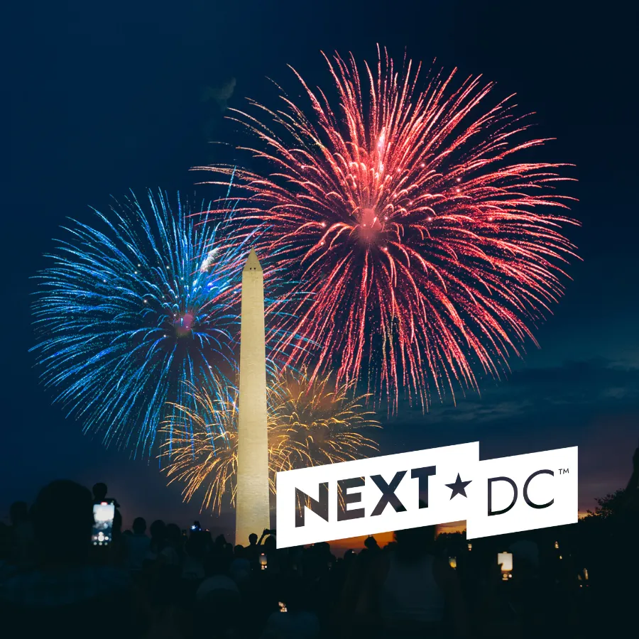 The Washington Monument with blue, gold, and red fireworks behind it. The NEXTDC logo in white is in the bottom corner.